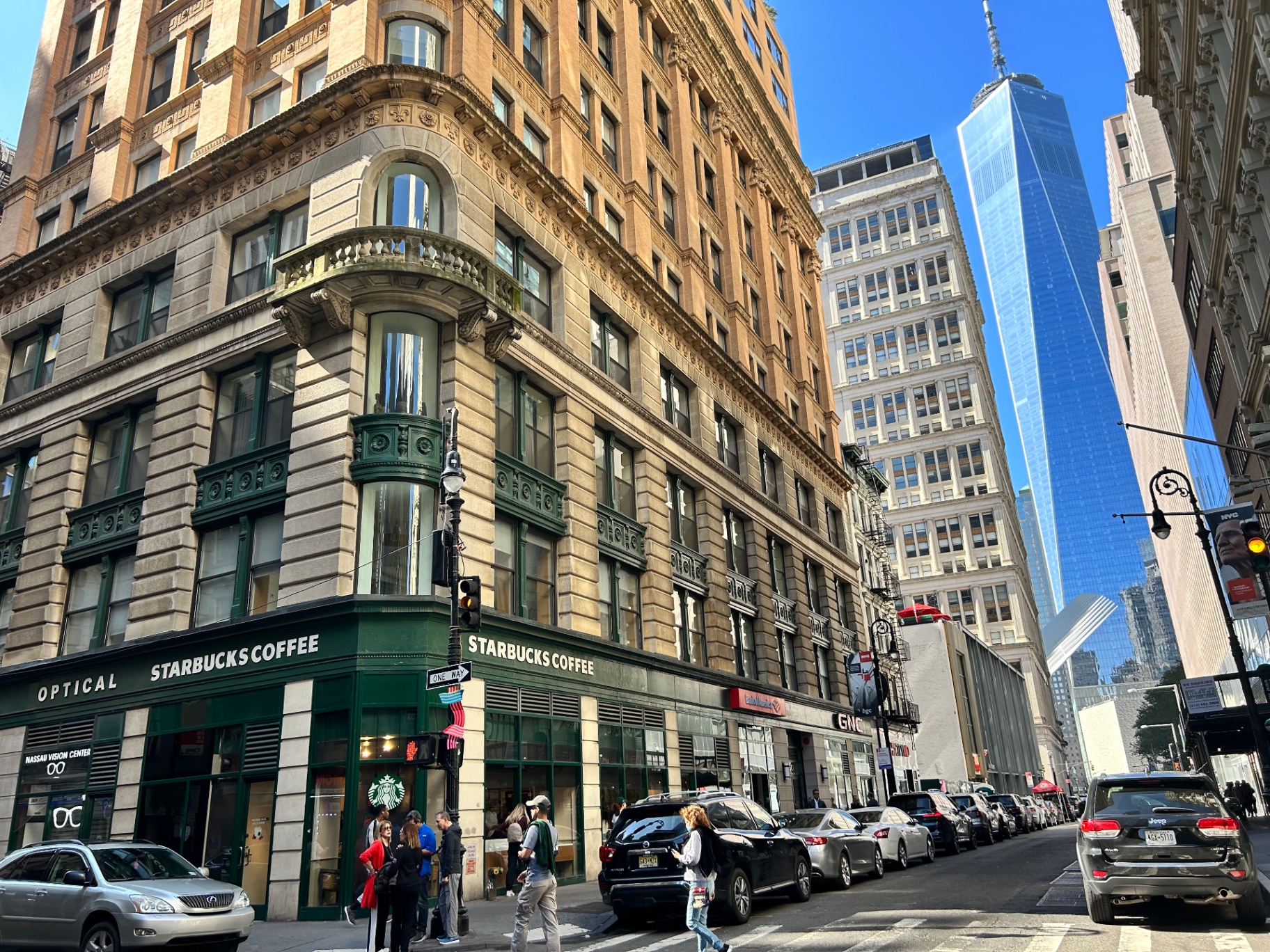 Lower Manhattan street corner with One World Trade Center towering in the background - classic NYC architecture meets modern skyline
